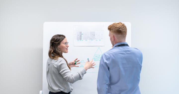 Two professionals discussing project plans at whiteboard in office setting.